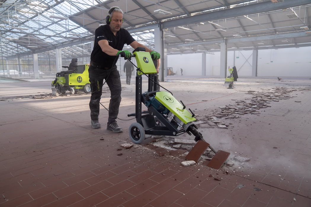 WOLFF_Application Images_RamHammer_Main_2022-11 Pracownik obsługuje zieloną elektryczną maszynę do usuwania podłóg, zrywając ceramiczne płytki w dużej, jasnej hali przemysłowej. Ma na sobie czarne polo, zielone robocze rękawice i ochronniki słuchu, popycha maszynę na kółkach przez połamane płytki i kurz. W tle widać inne urządzenia i pracowników pod wysokim szklanym dachem; gruz i odczepione płytki pokrywają posadzkę. Scena przedstawia trwające prace remontowe podłogi z pyłem, kablami i wyposażeniem ochronnym.