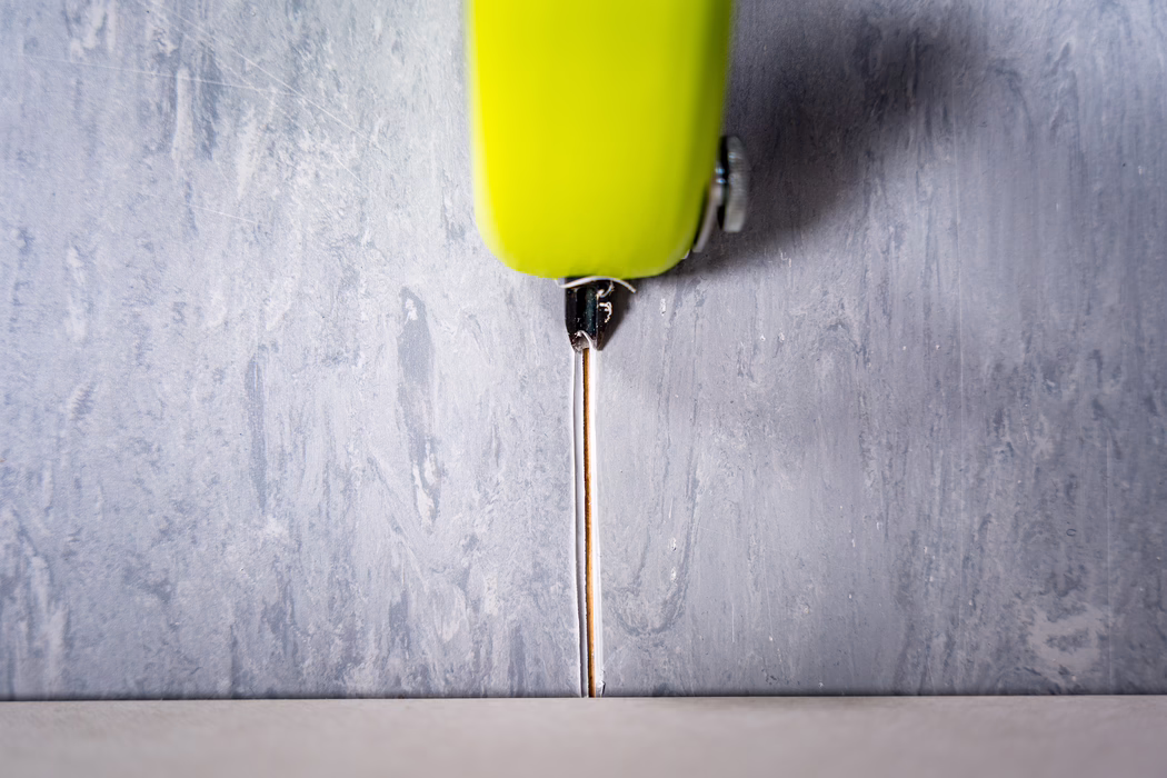Zbliżenie limonkowo-zielonego chowanego nożyka ustawionego pionowo nad wąską fugą między fakturowaną szarą ścianą a jasną podłogą. Cienkie metalowe ostrze jest wysunięte w dół w fugę, z plastikowym uchwytem i małą metalową śrubą widocznymi u góry kadru. Ujęcie podkreśla kontrastujące tekstury — szorstki, malowany tynk na ścianie i gładką powierzchnię podłogi — i jest delikatnie oświetlone z góry, co uwydatnia drobne zadrapania i ślady zużycia na ostrzu i uchwycie.