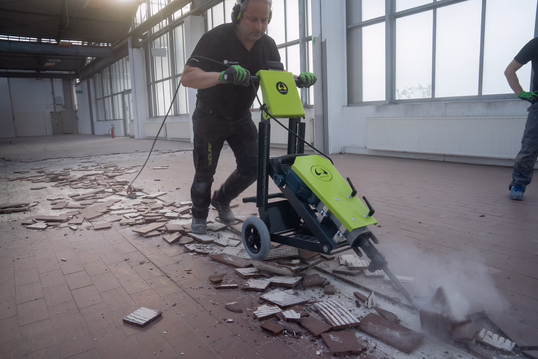 WOLFF_Application Images_RamHammer_Detail3_2022-11 A construction worker wearing protective gloves and earmuffs operates a bright green electric floor scraper to remove ceramic tiles inside a large, empty industrial hall. Broken tiles and dust are scattered across the reddish tile floor while the machine lifts and fractures the tile layer, producing white dust and debris. The worker pushes the wheeled machine forward, firmly holding the handle and guiding the attached chisel, while a second person is partially visible at the right edge of the frame. Tall industrial windows on the right provide diffuse daylight, and the high ceiling with steel beams emphasizes the scale of the renovation work in progress. The scene highlights action, tools, safety equipment and the process of removing old flooring.