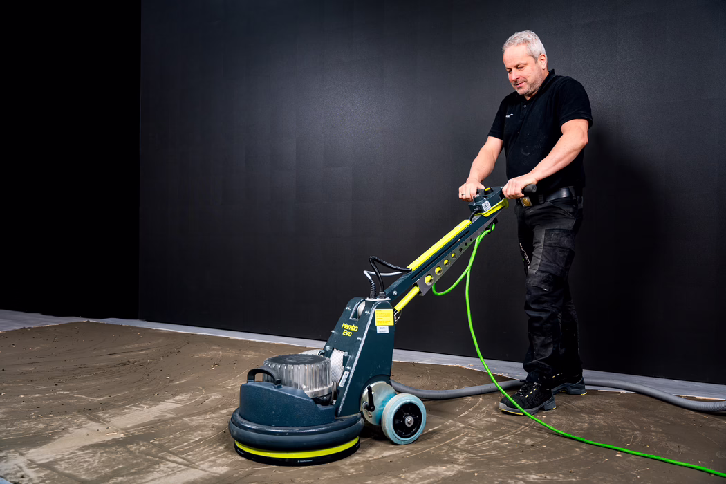 WOLFF_Application Image_Mambo_2024-01 (17) A man in black work clothes operates a professional floor polishing machine indoors. He stands at the right side of the frame, holding the handle of a green-and-gray rotary floor machine labeled 'Mambo EVO' and guiding it across a rough, partially smoothed concrete or screed floor with visible tool marks, dust and debris. A bright green power cable and a thick grey hose trail from the machine across the floor; a large dark panel forms the neutral background, giving the scene a studio-like, well-lit appearance that highlights floor texture and the worker's focused posture. The composition emphasizes the equipment, the work surface and the ongoing floor preparation task in a professional renovation setting.